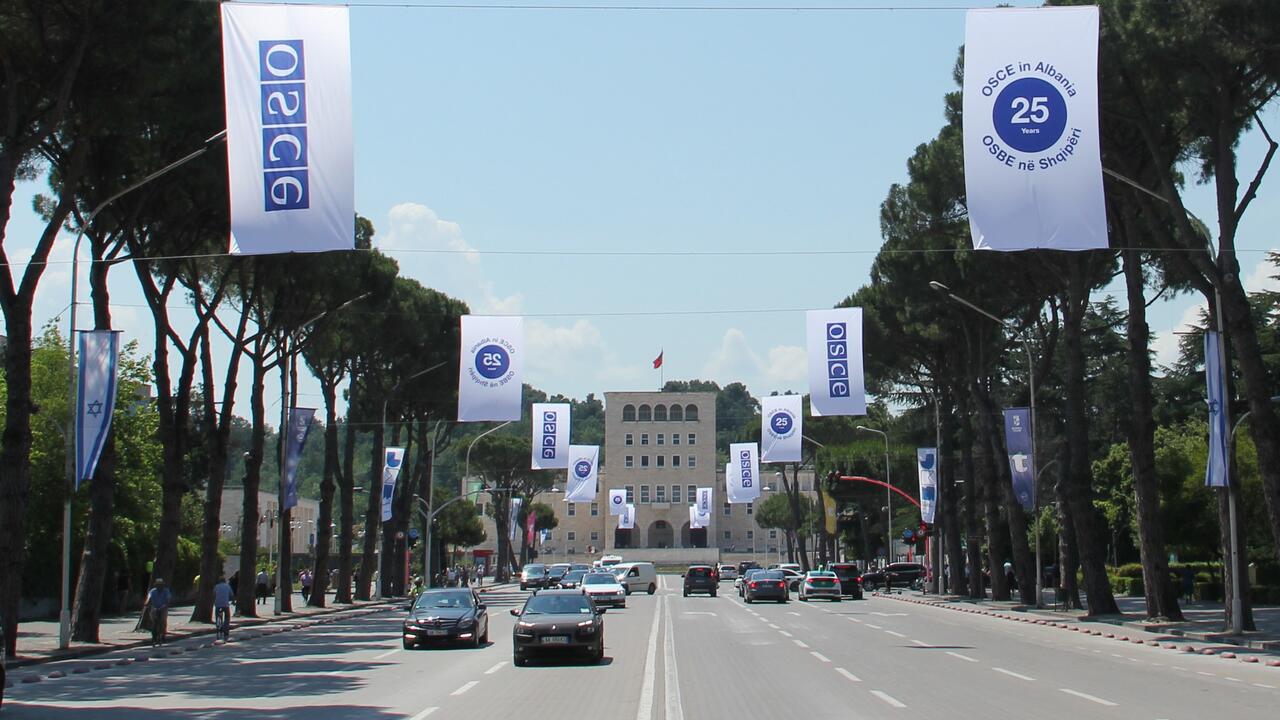 Wide road flanked by trees and banners, leading to a distant building under a clear blue sky.